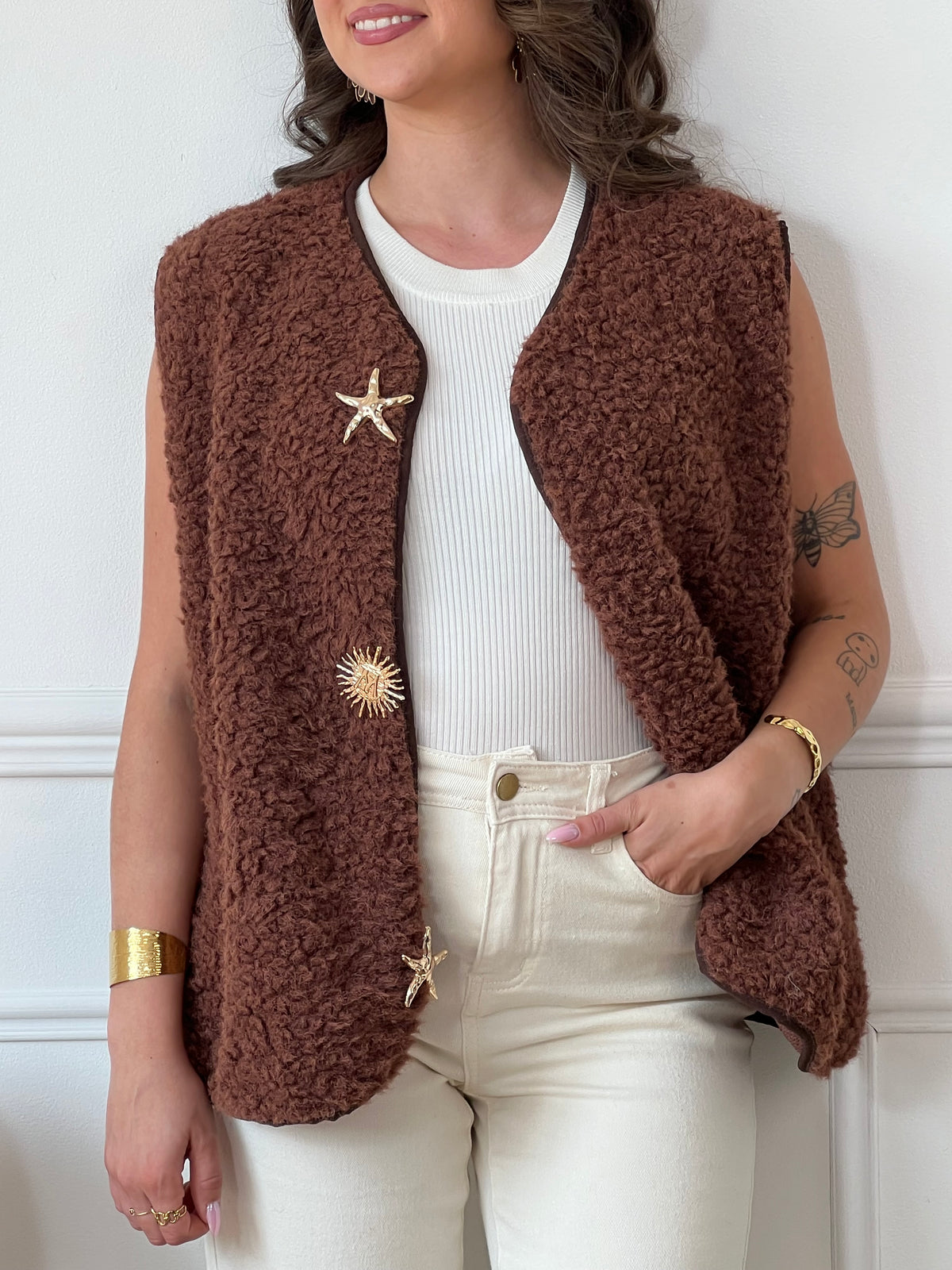La veste moumoute sans manches Eda allie douceur et style. Conçue pour offrir un confort optimal, elle présente un col en V élégant et des broches dorées délicates sur le devant, qui ajoutent une touche sophistiquée.