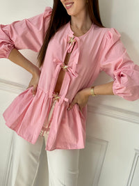 Cette blouse rose Adalia allie élégance et confort avec ses manches longues bouffantes, son col rond et ses poignets élastiques. Les liens à nouer sur le devant et les volants délicats sur le bas apportent une touche féminine et raffinée, idéale pour sublimer votre style au quotidien.