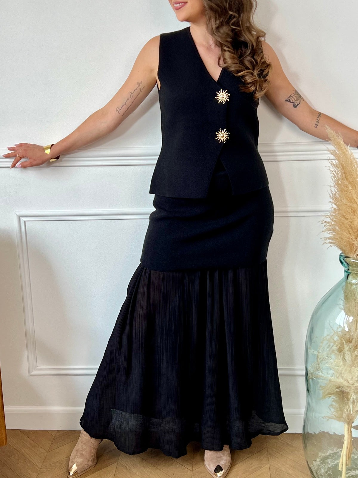 Ce gilet sans manches noir, nommé Elvire, allie élégance et originalité. Sa fermeture à boutons sur toute la longueur est sublimée par un détail doré en forme de soleil, apportant une touche lumineuse à votre tenue. Son col en V flatteur met en valeur votre silhouette, tandis que sa coupe sans manches offre confort et liberté de mouvement.