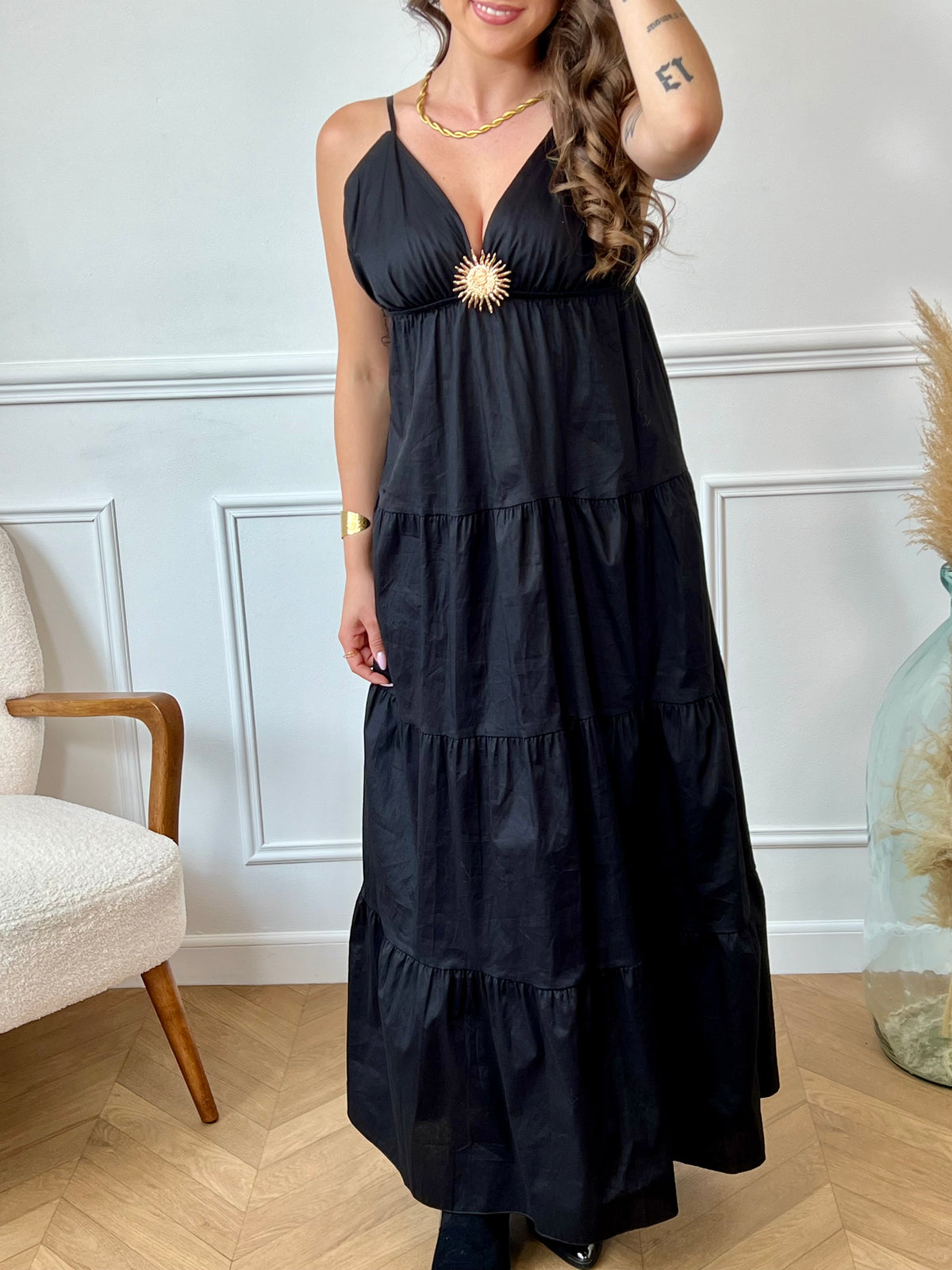 Cette robe longue noire, Elenora, allie élégance et confort grâce à ses bretelles réglables et sa taille élastique qui s’adaptent parfaitement à votre silhouette. Son col en V met en valeur le décolleté, tandis que le détail doré en forme de soleil apporte une touche lumineuse et raffinée sur la poitrine.
