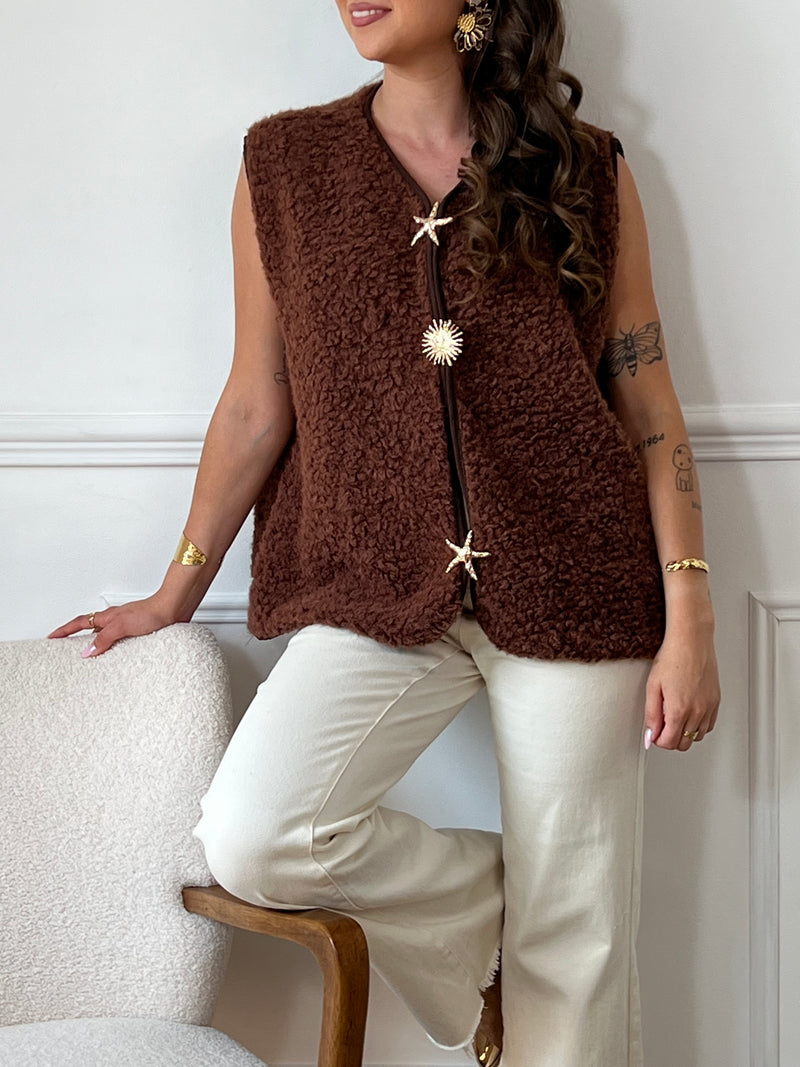 La veste moumoute sans manches Eda allie douceur et style. Conçue pour offrir un confort optimal, elle présente un col en V élégant et des broches dorées délicates sur le devant, qui ajoutent une touche sophistiquée.