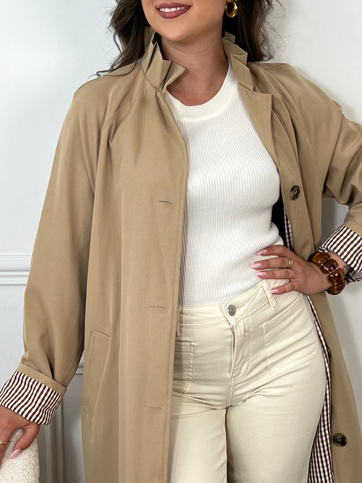 Trench beige femme avec ceinture, manches longues et col volanté pour une touche féminine. Boutons tout le long, doublure intérieure en carreaux vichy marron et écru, offrant élégance et confort. Coupe ample et deux poches latérales pratiques.