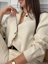 Ce gilet beige Eloisa allie élégance et féminité avec ses manches longues et son col rond délicatement agrémenté de volants. La ligne de boutons noirs, ornés de détails dorés et d’une perle, apporte une touche raffinée qui souligne subtilement la silhouette.