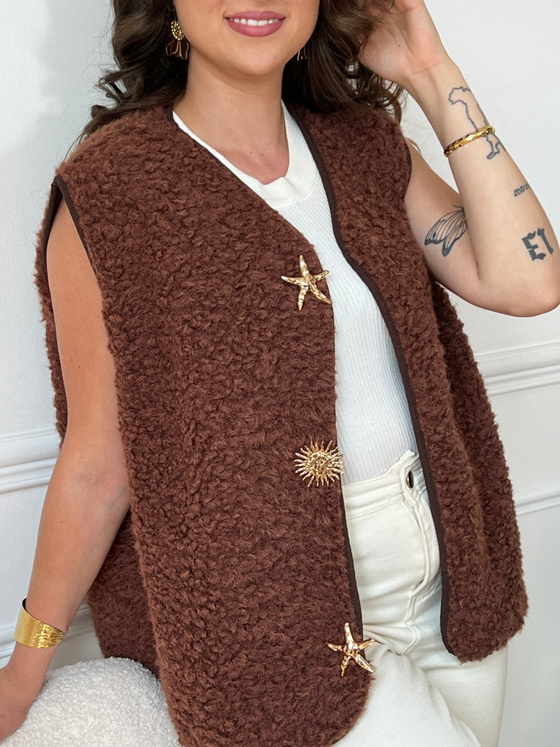 La veste moumoute sans manches Eda allie douceur et style. Conçue pour offrir un confort optimal, elle présente un col en V élégant et des broches dorées délicates sur le devant, qui ajoutent une touche sophistiquée.
