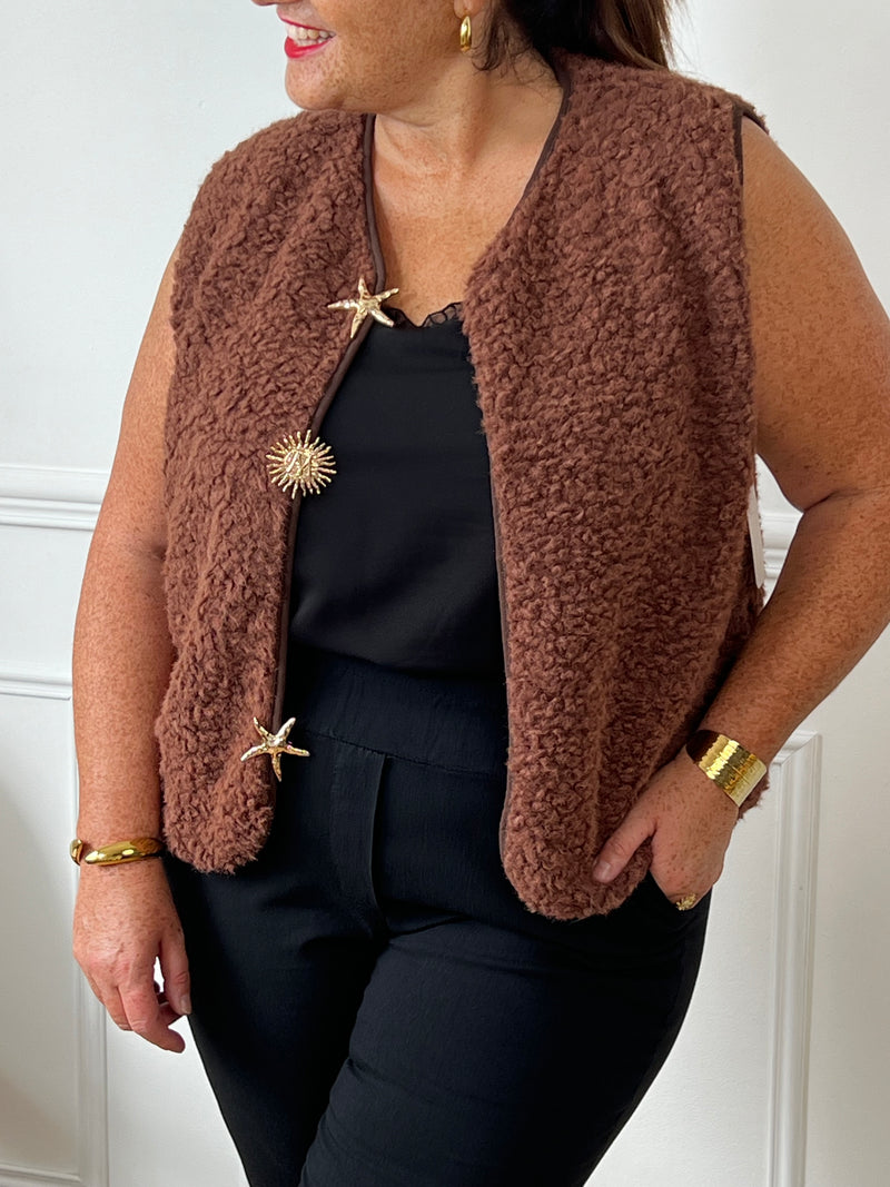 La veste moumoute sans manches Eda allie douceur et style. Conçue pour offrir un confort optimal, elle présente un col en V élégant et des broches dorées délicates sur le devant, qui ajoutent une touche sophistiquée.