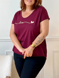 Tee-shirt bordeaux grande taille pour femme, manches courtes et col V élégant. Sa coupe moderne plus longue à l’arrière offre confort et style. Détail chic : inscription dorée "Love". Couleurs tendance bordeaux et doré, idéal pour un look casual et féminin