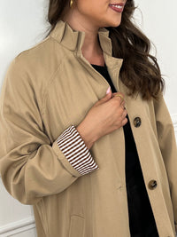 Trench beige femme avec ceinture, manches longues et col volanté pour une touche féminine. Boutons tout le long, doublure intérieure en carreaux vichy marron et écru, offrant élégance et confort. Coupe ample et deux poches latérales pratiques.