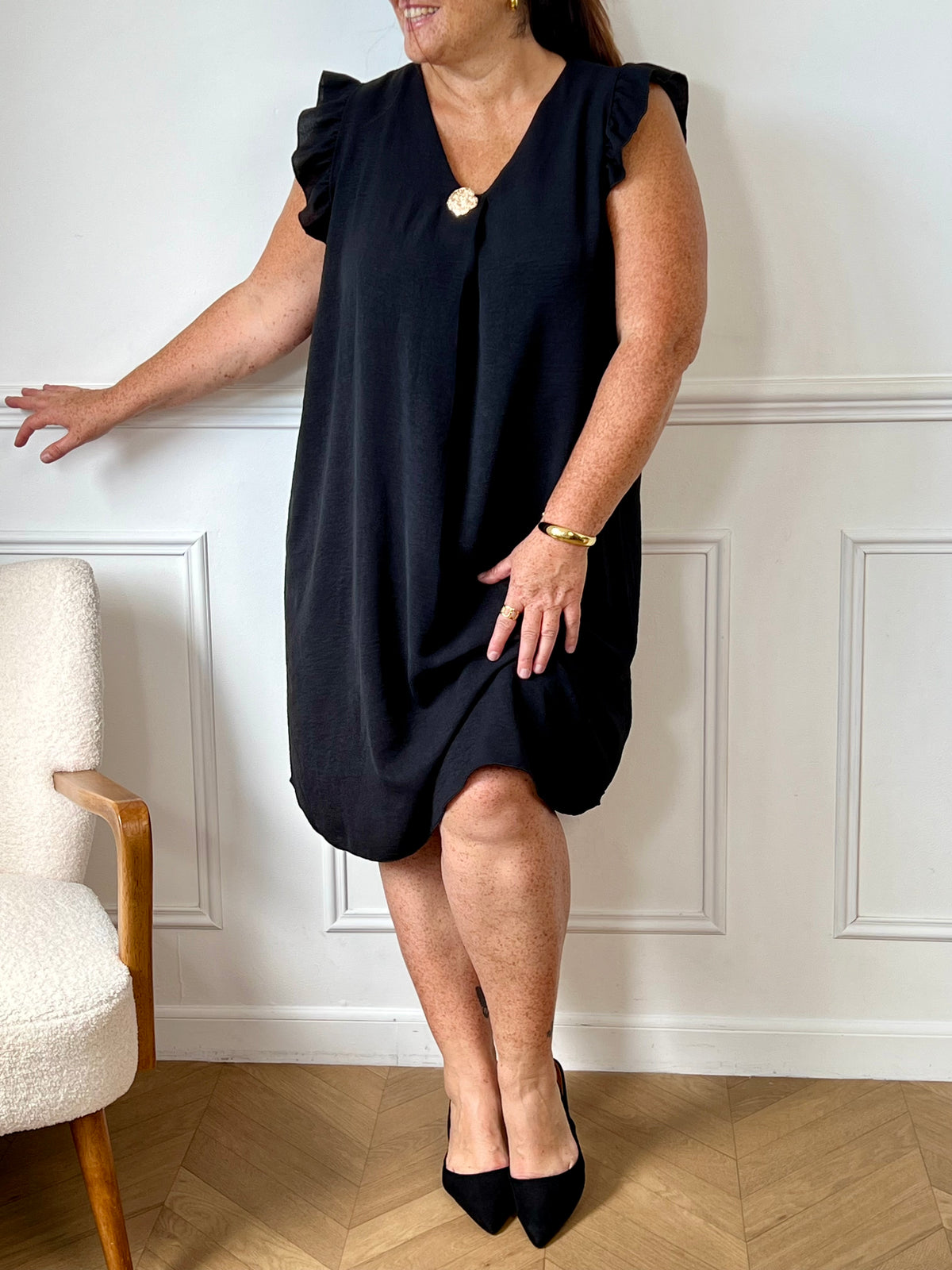 Cette robe noire sans manches, parfaite pour les grandes tailles, offre une silhouette élégante avec ses volants délicats aux épaules. Le col en V met en valeur le décolleté tandis que l'épingle dorée sur le devant ajoute une touche raffinée.