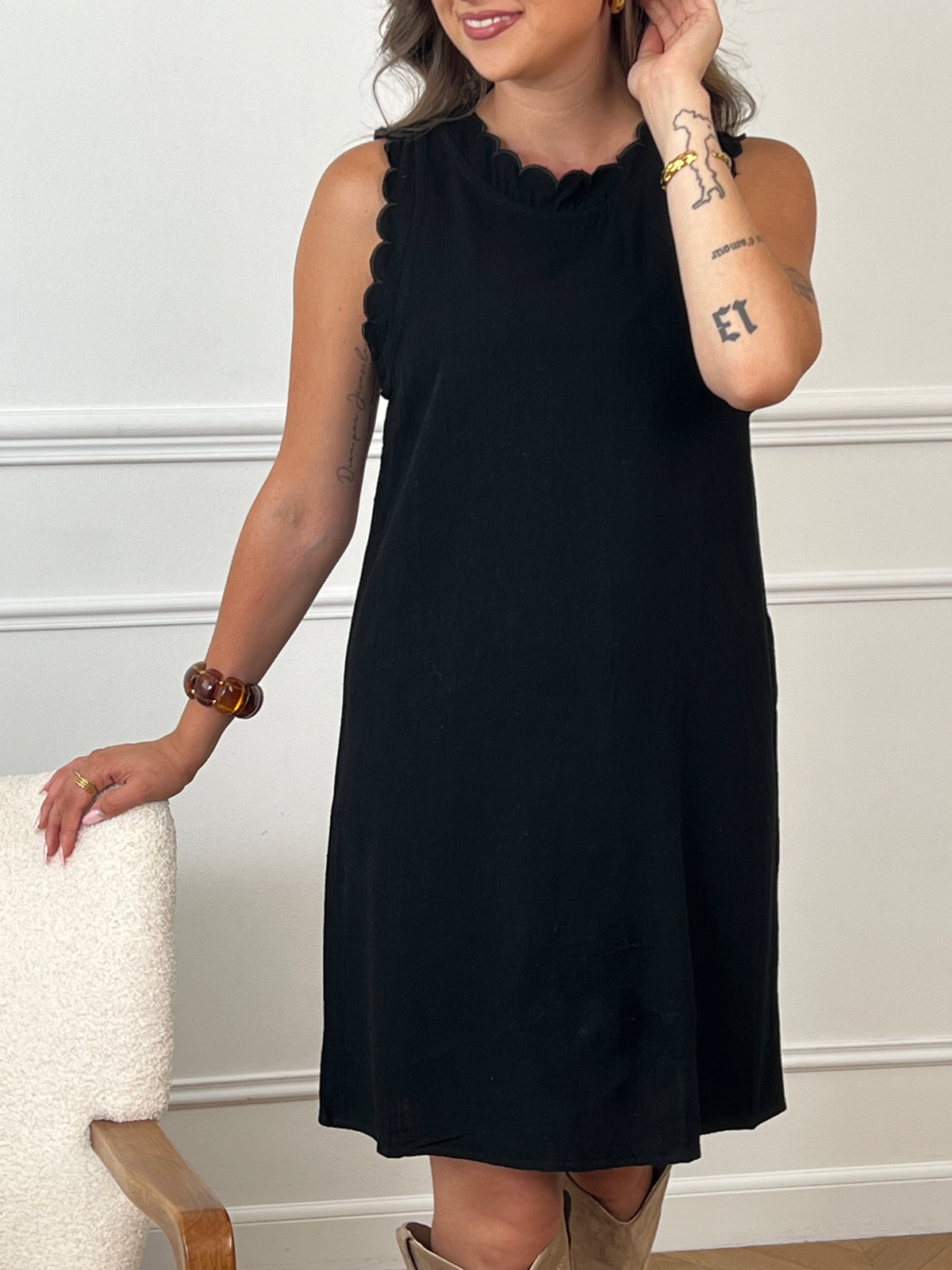 La robe courte noire Floriane est sans manches, avec un col rond devant et derrière. Ses détails festonnés et son ouverture dos avec lien à nouer ajoutent une touche délicate et féminine.
