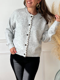 Ce gilet gris Eloisa allie élégance et féminité avec ses manches longues et son col rond délicatement agrémenté de volants. La ligne de boutons noirs, ornés de détails dorés et d’une perle, apporte une touche raffinée qui souligne subtilement la silhouette.