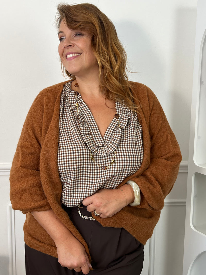 Blouse Nour à petits carreaux marron et écru, avec encolure volantée, portée sous un gilet camel.