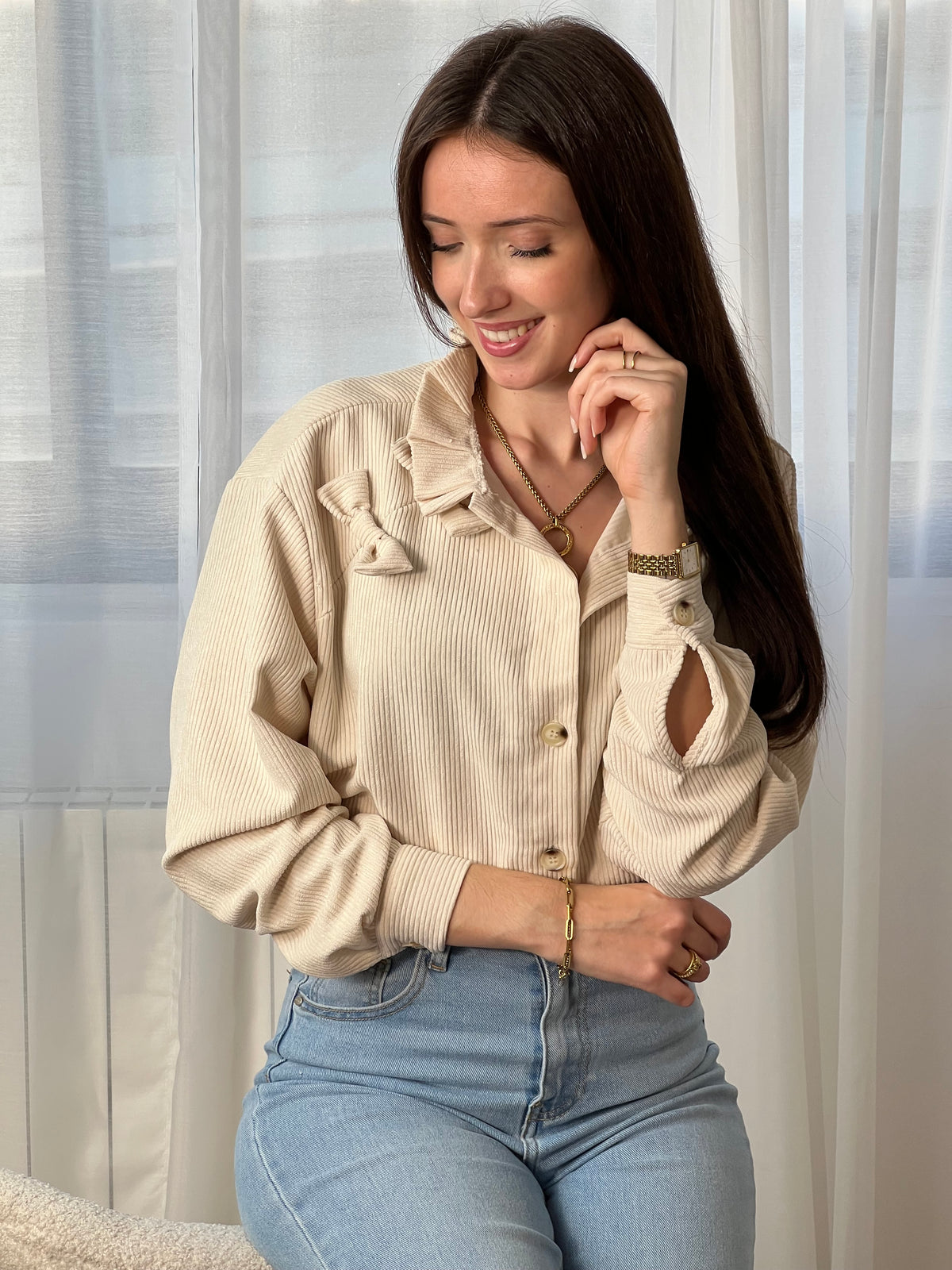 Chemise beige en velours côtelé pour femme, dotée de manches longues et d'un col rond orné de volants. Nœuds latéraux et boutons discrets sur toute la longueur et aux poignets pour un style raffiné et élégant.