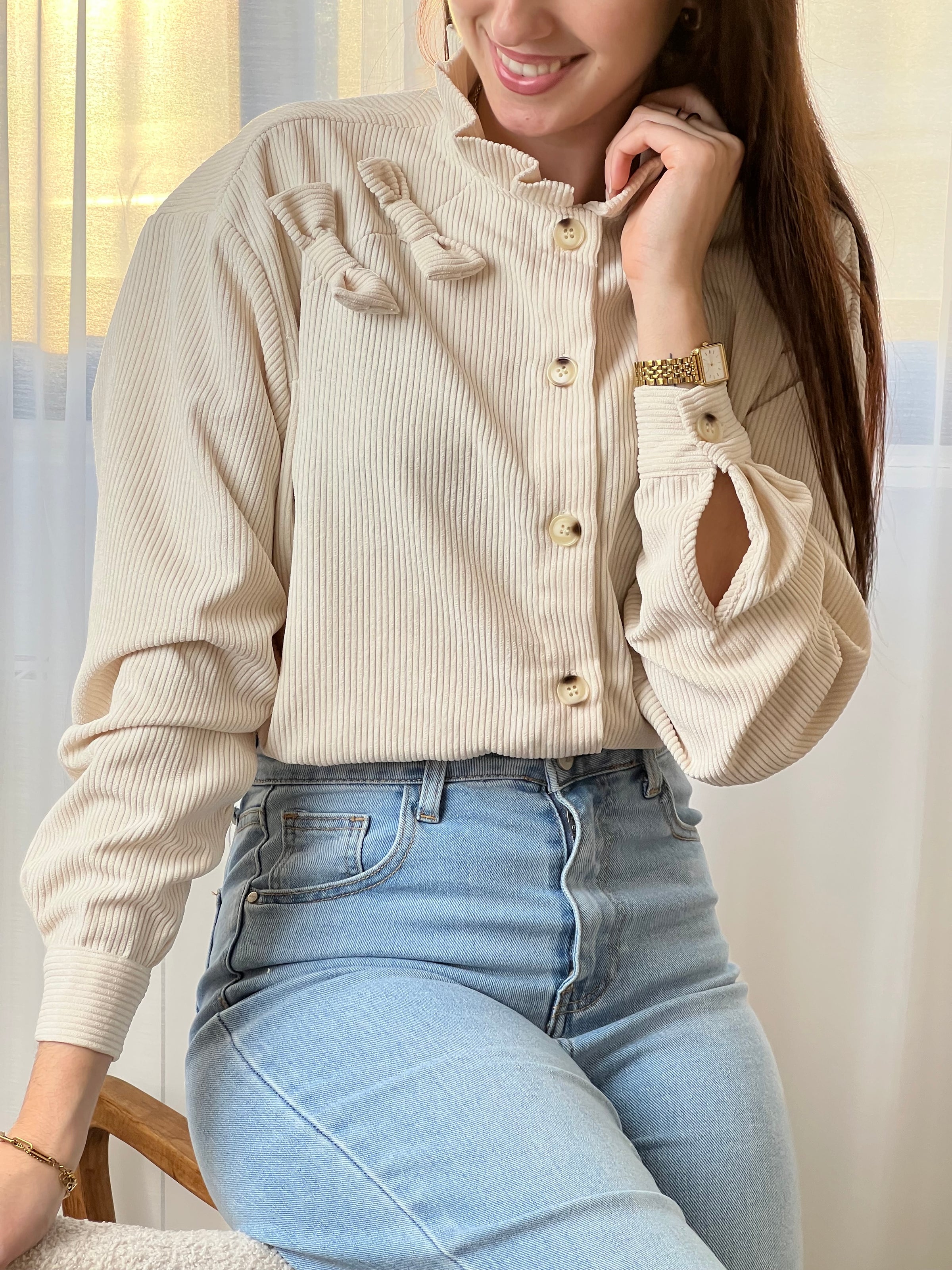 Chemise beige en velours côtelé pour femme, dotée de manches longues et d'un col rond orné de volants. Nœuds latéraux et boutons discrets sur toute la longueur et aux poignets pour un style raffiné et élégant.