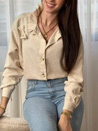 Chemise beige en velours côtelé pour femme, dotée de manches longues et d'un col rond orné de volants. Nœuds latéraux et boutons discrets sur toute la longueur et aux poignets pour un style raffiné et élégant.