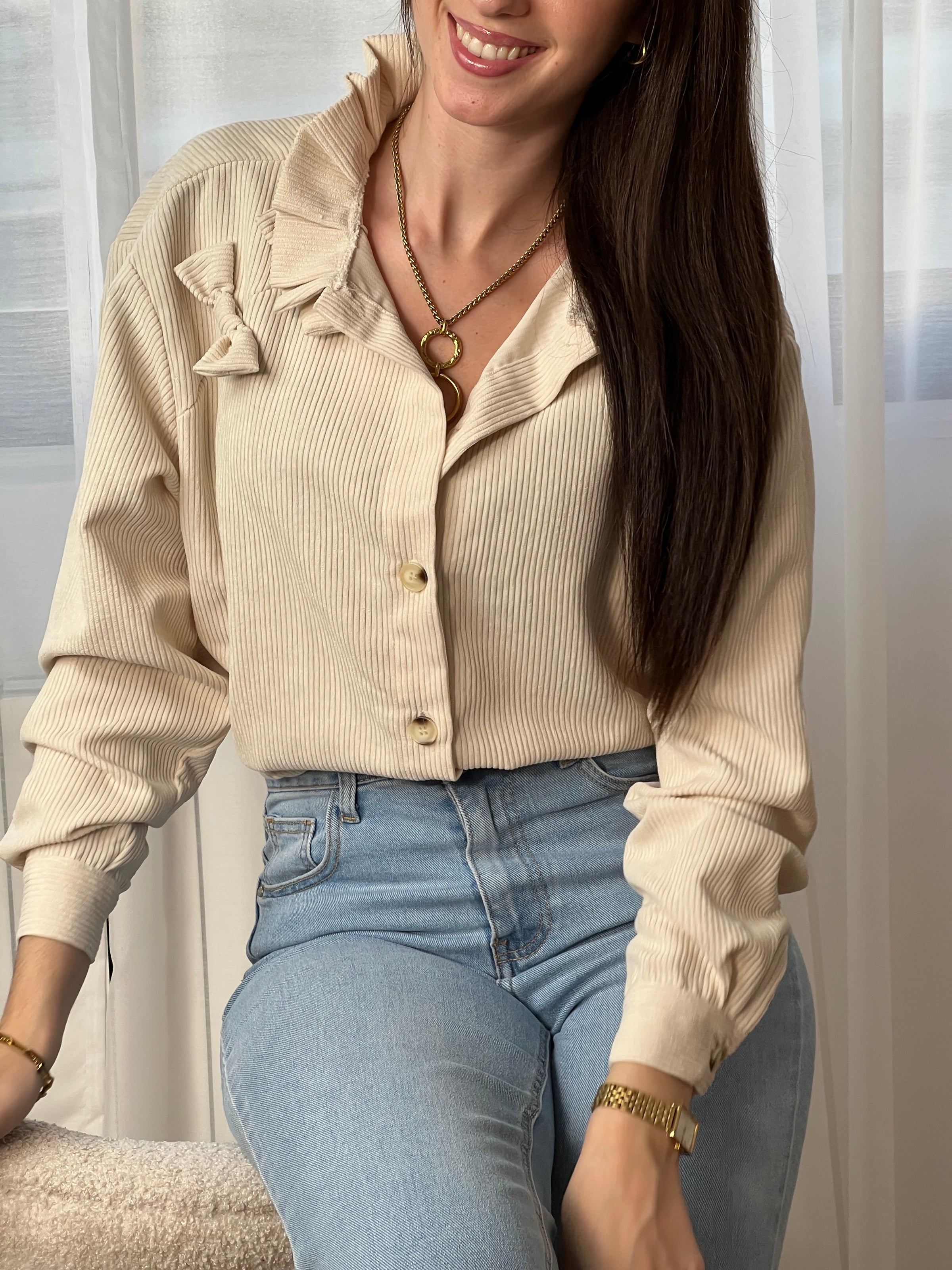 Chemise beige en velours côtelé pour femme, dotée de manches longues et d'un col rond orné de volants. Nœuds latéraux et boutons discrets sur toute la longueur et aux poignets pour un style raffiné et élégant.