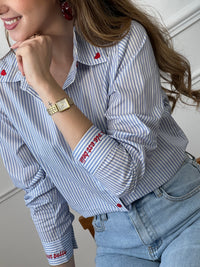 Chemise en coton femme, parfaite pour un look à la fois élégant et original. Dotée de manches longues, d’un col chemise et de boutons rouges sur toute la longueur ainsi qu’aux poignets, elle se distingue par ses rayures verticales bleues et écrues. Ses détails brodés, avec des cœurs au col et l’inscription "La vie est belle" aux poignets, apportent une touche unique et tendance