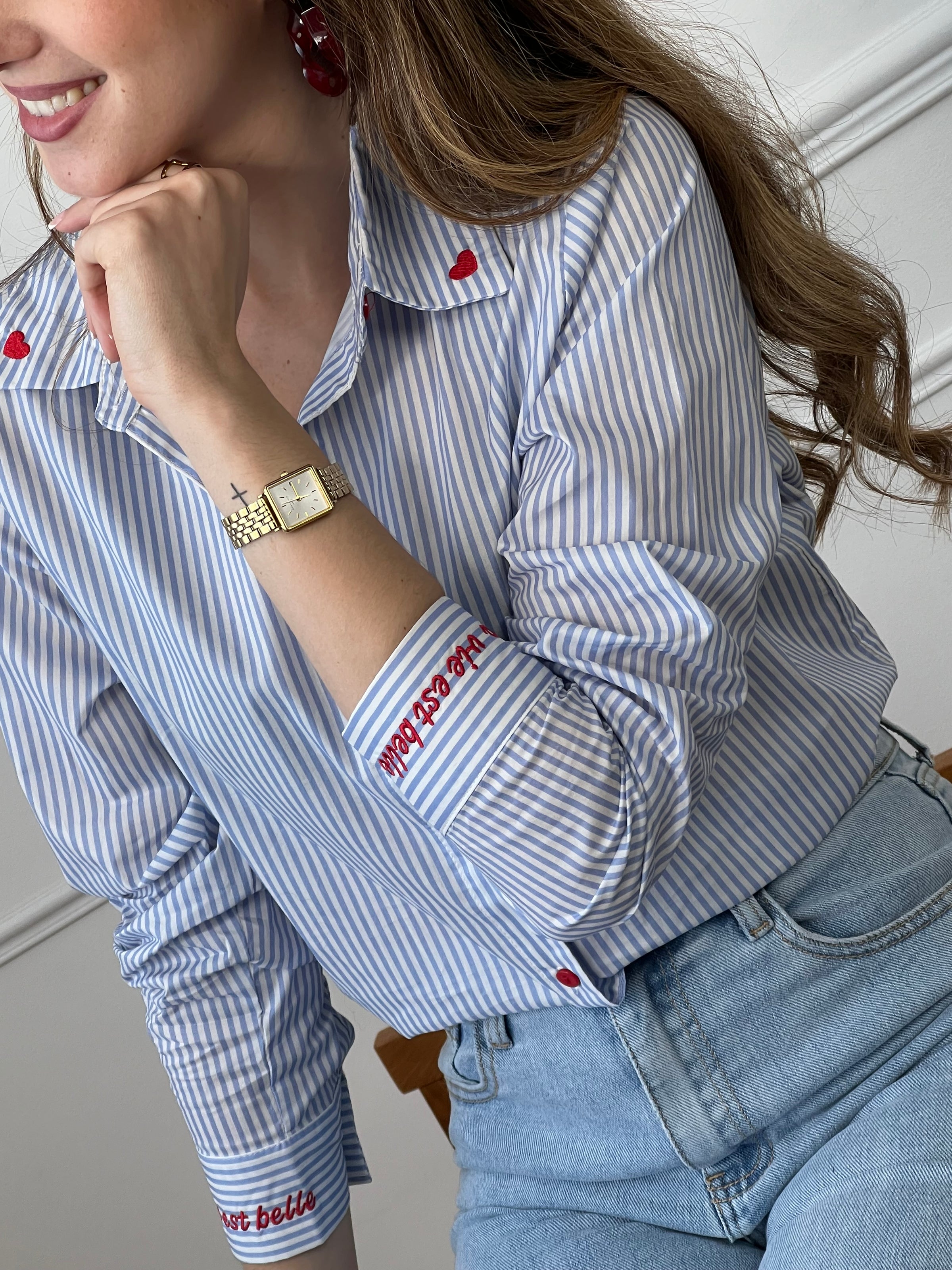 Chemise en coton femme, parfaite pour un look à la fois élégant et original. Dotée de manches longues, d’un col chemise et de boutons rouges sur toute la longueur ainsi qu’aux poignets, elle se distingue par ses rayures verticales bleues et écrues. Ses détails brodés, avec des cœurs au col et l’inscription "La vie est belle" aux poignets, apportent une touche unique et tendance