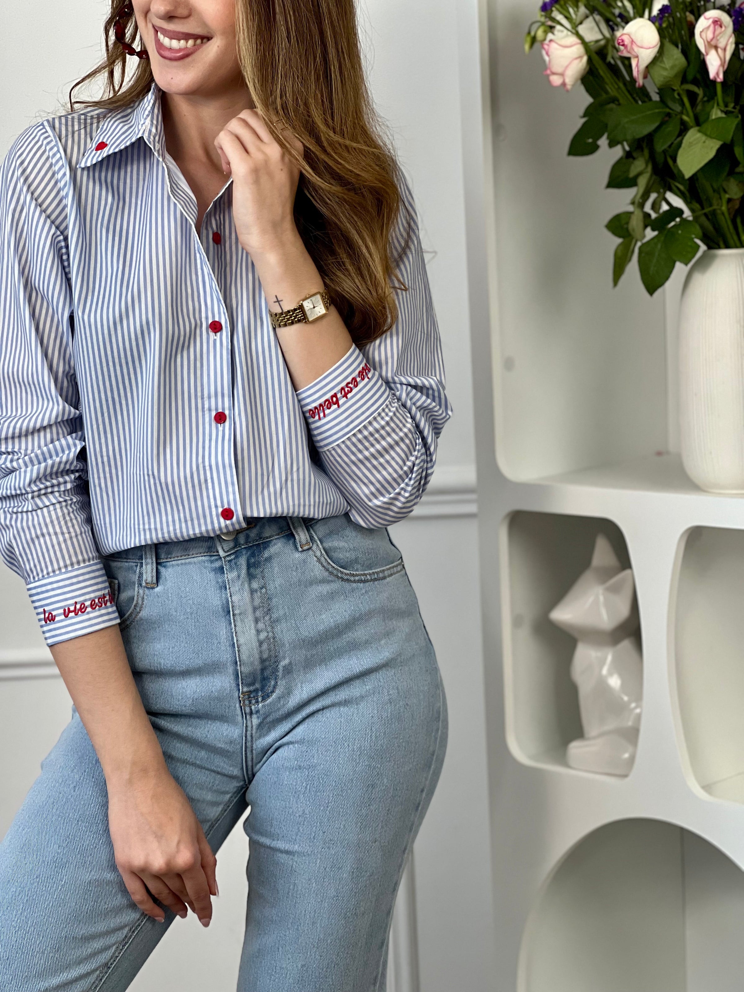 Chemise en coton femme, parfaite pour un look à la fois élégant et original. Dotée de manches longues, d’un col chemise et de boutons rouges sur toute la longueur ainsi qu’aux poignets, elle se distingue par ses rayures verticales bleues et écrues. Ses détails brodés, avec des cœurs au col et l’inscription "La vie est belle" aux poignets, apportent une touche unique et tendance