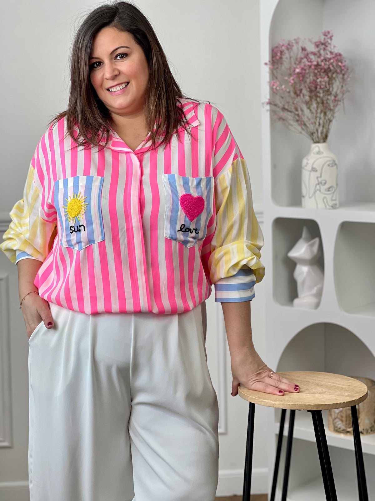 Chemise grande taille colorée au style oversize, idéale pour un look tendance et décontracté. Avec sa coupe longue, légèrement plus étendue au dos, ses manches longues et son col chemise classique, elle allie confort et modernité. Dotée de boutons sur toute la longueur, de poignets boutonnés et de deux poches poitrine, elle se distingue par ses rayures roses, jaunes, bleues et écrues ainsi que ses broderies originales "sun", "love", cœur et soleil.