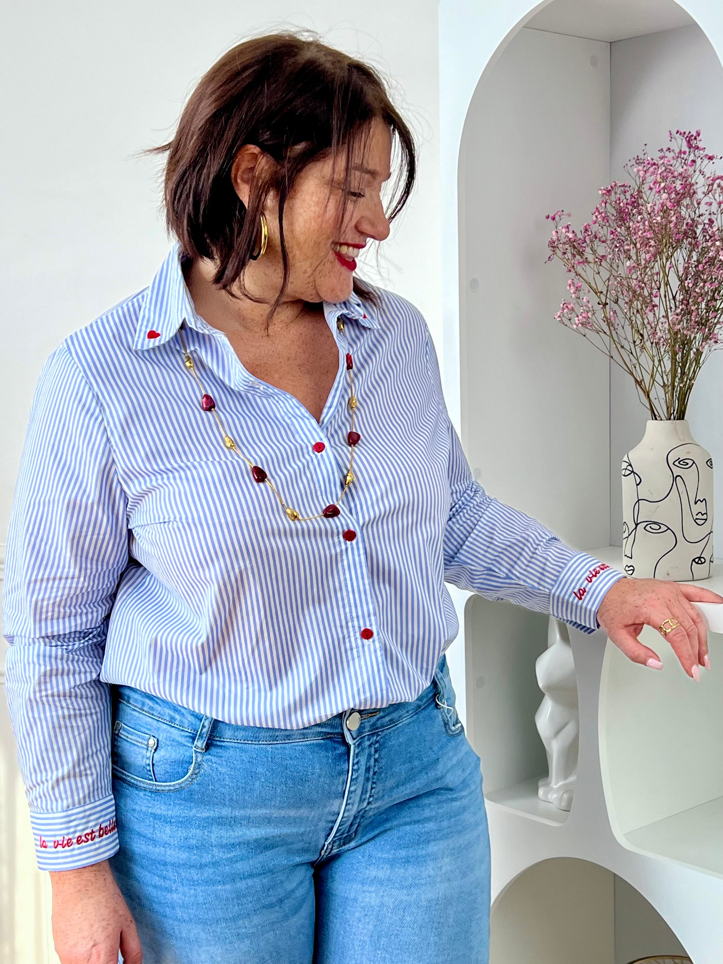 Chemise en coton grande taille femme, parfaite pour un look à la fois élégant et original. Dotée de manches longues, d’un col chemise et de boutons rouges sur toute la longueur ainsi qu’aux poignets, elle se distingue par ses rayures verticales bleues et écrues. Ses détails brodés, avec des cœurs au col et l’inscription "La vie est belle" aux poignets, apportent une touche unique et tendance