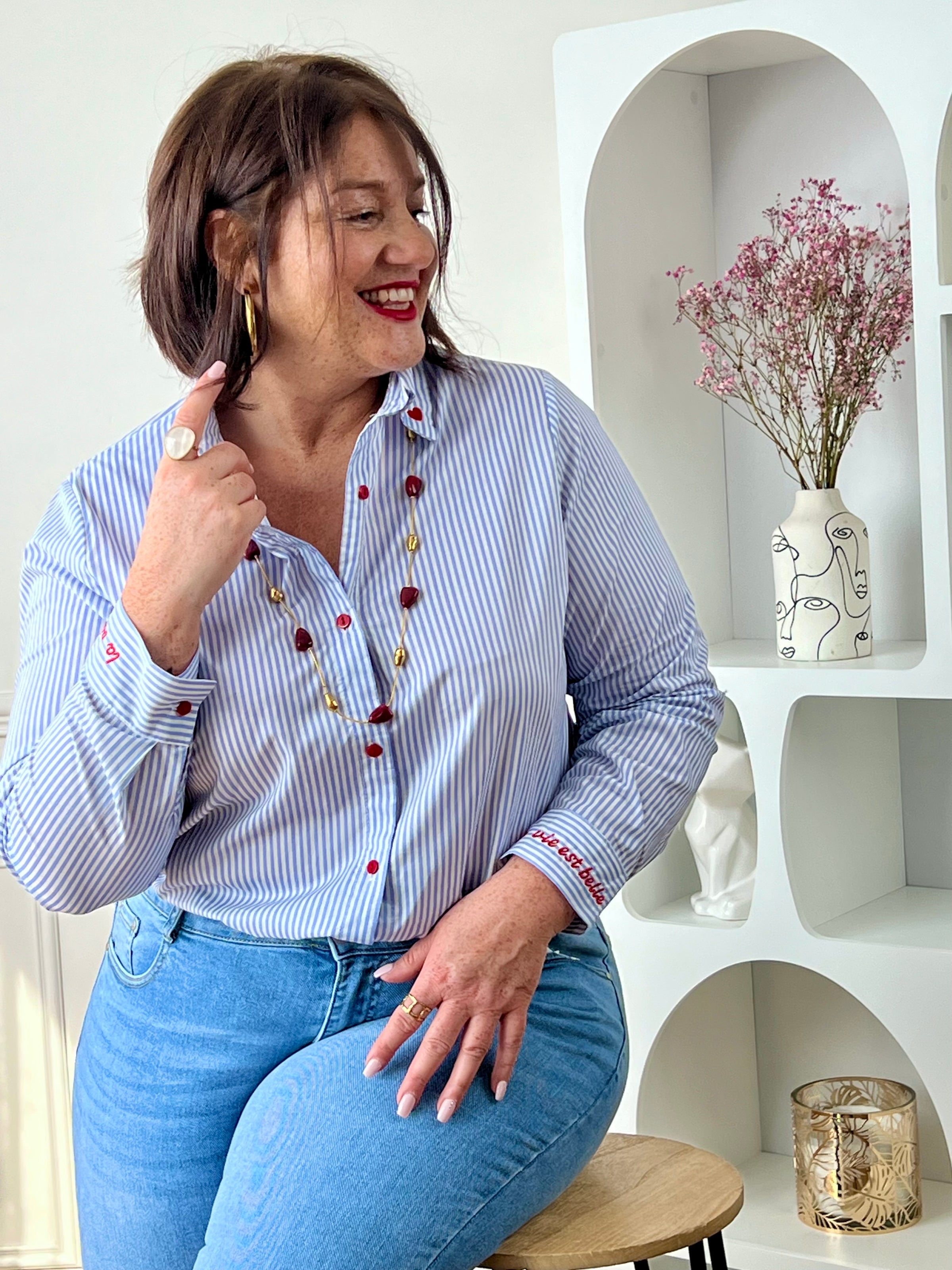 Chemise en coton grande taille femme, parfaite pour un look à la fois élégant et original. Dotée de manches longues, d’un col chemise et de boutons rouges sur toute la longueur ainsi qu’aux poignets, elle se distingue par ses rayures verticales bleues et écrues. Ses détails brodés, avec des cœurs au col et l’inscription "La vie est belle" aux poignets, apportent une touche unique et tendance