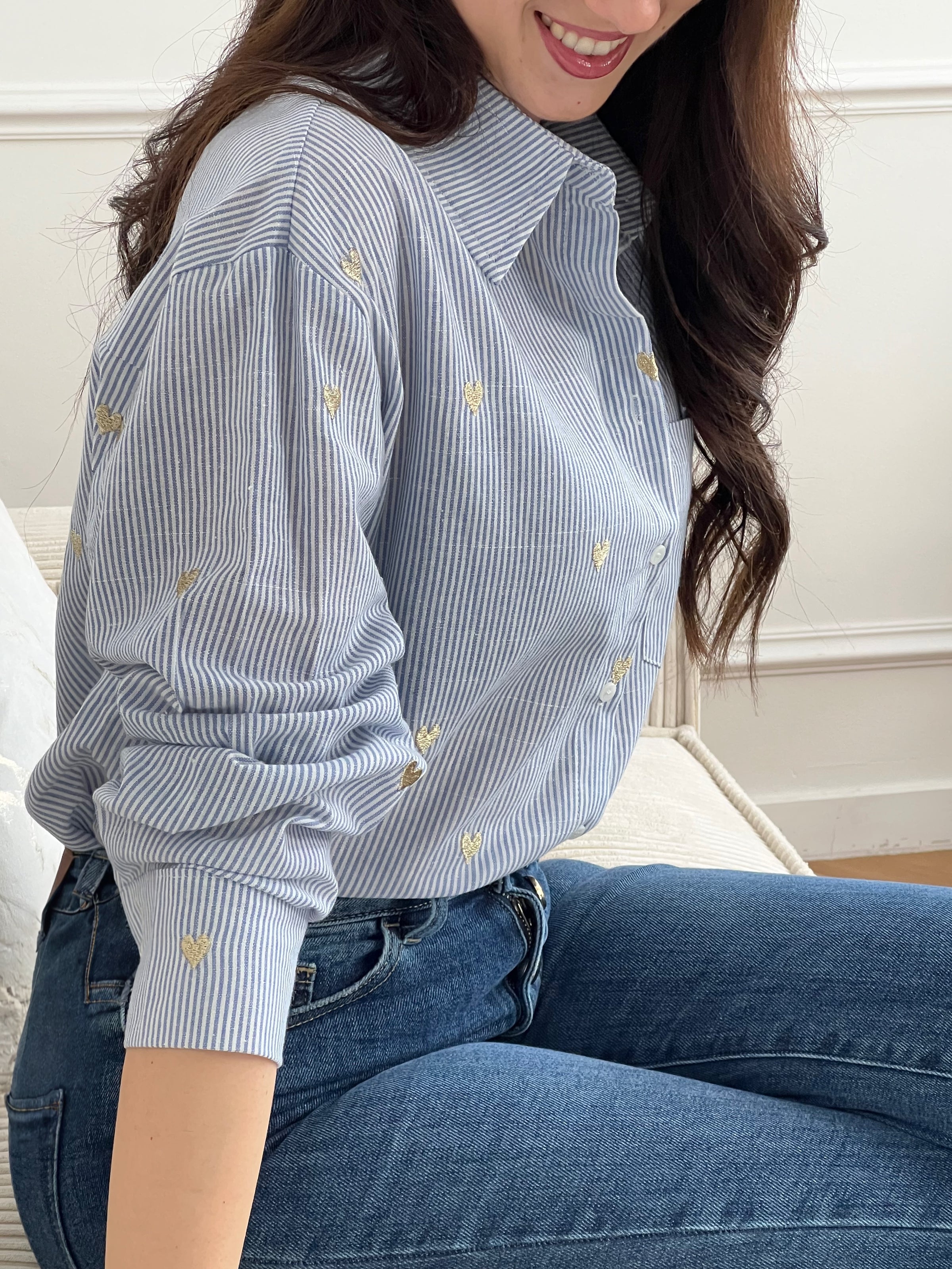 La chemise Sélène en coton combine style intemporel et détails délicats. Rayures verticales bleu et écru, cœurs dorés brodés et col chemise structuré créent une allure sophistiquée. Manches longues avec boutons aux poignets, poche poitrine et fermeture boutonnée complète.