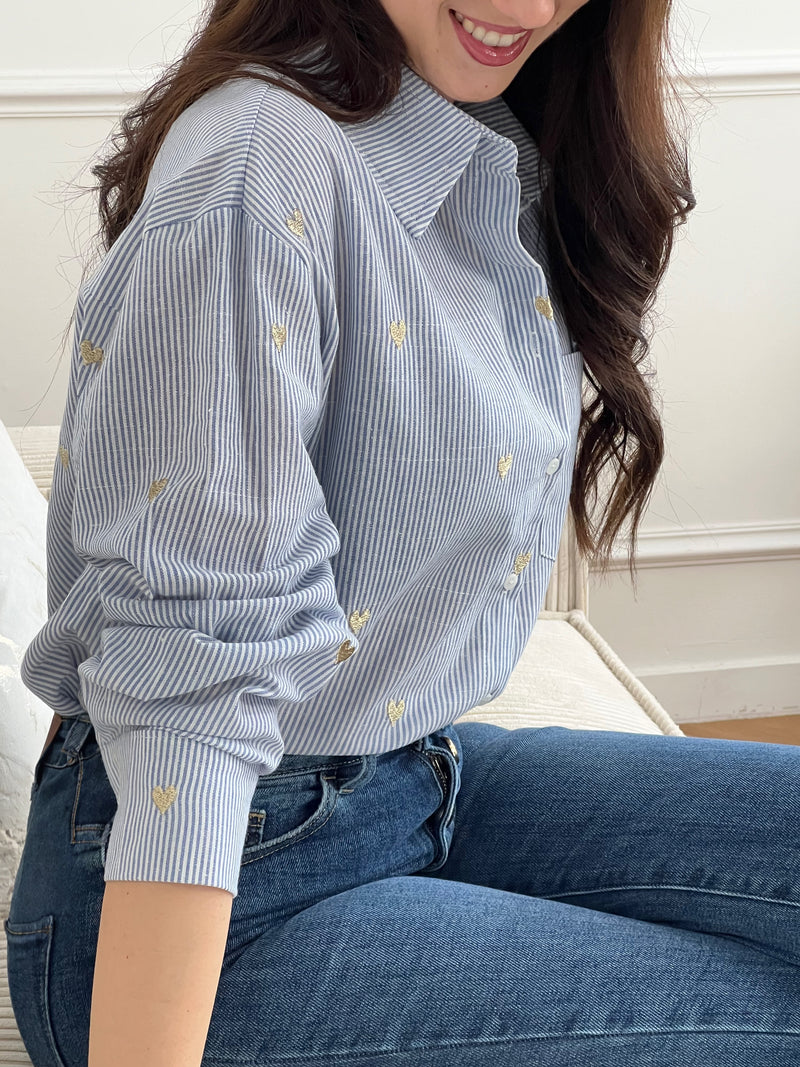 La chemise Sélène en coton combine style intemporel et détails délicats. Rayures verticales bleu et écru, cœurs dorés brodés et col chemise structuré créent une allure sophistiquée. Manches longues avec boutons aux poignets, poche poitrine et fermeture boutonnée complète.