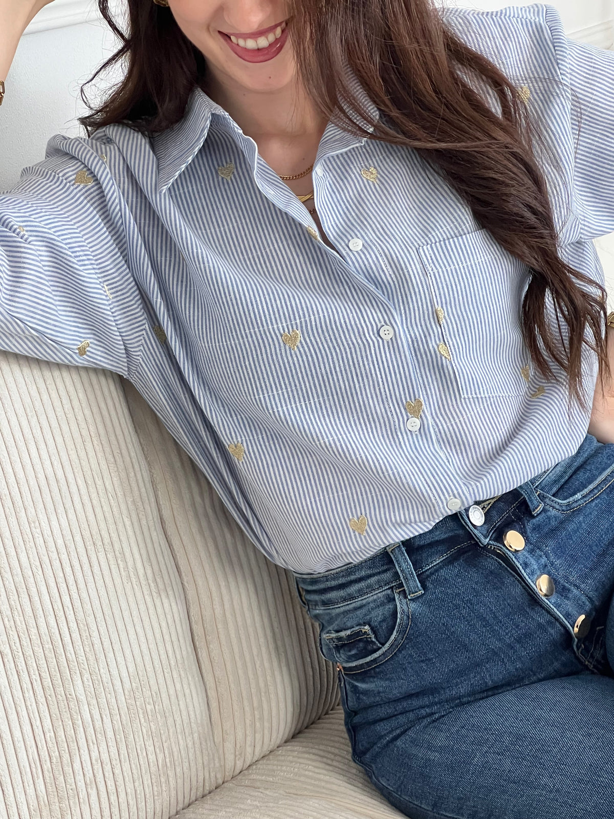 La chemise Sélène en coton combine style intemporel et détails délicats. Rayures verticales bleu et écru, cœurs dorés brodés et col chemise structuré créent une allure sophistiquée. Manches longues avec boutons aux poignets, poche poitrine et fermeture boutonnée complète.