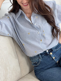 La chemise Sélène en coton combine style intemporel et détails délicats. Rayures verticales bleu et écru, cœurs dorés brodés et col chemise structuré créent une allure sophistiquée. Manches longues avec boutons aux poignets, poche poitrine et fermeture boutonnée complète.