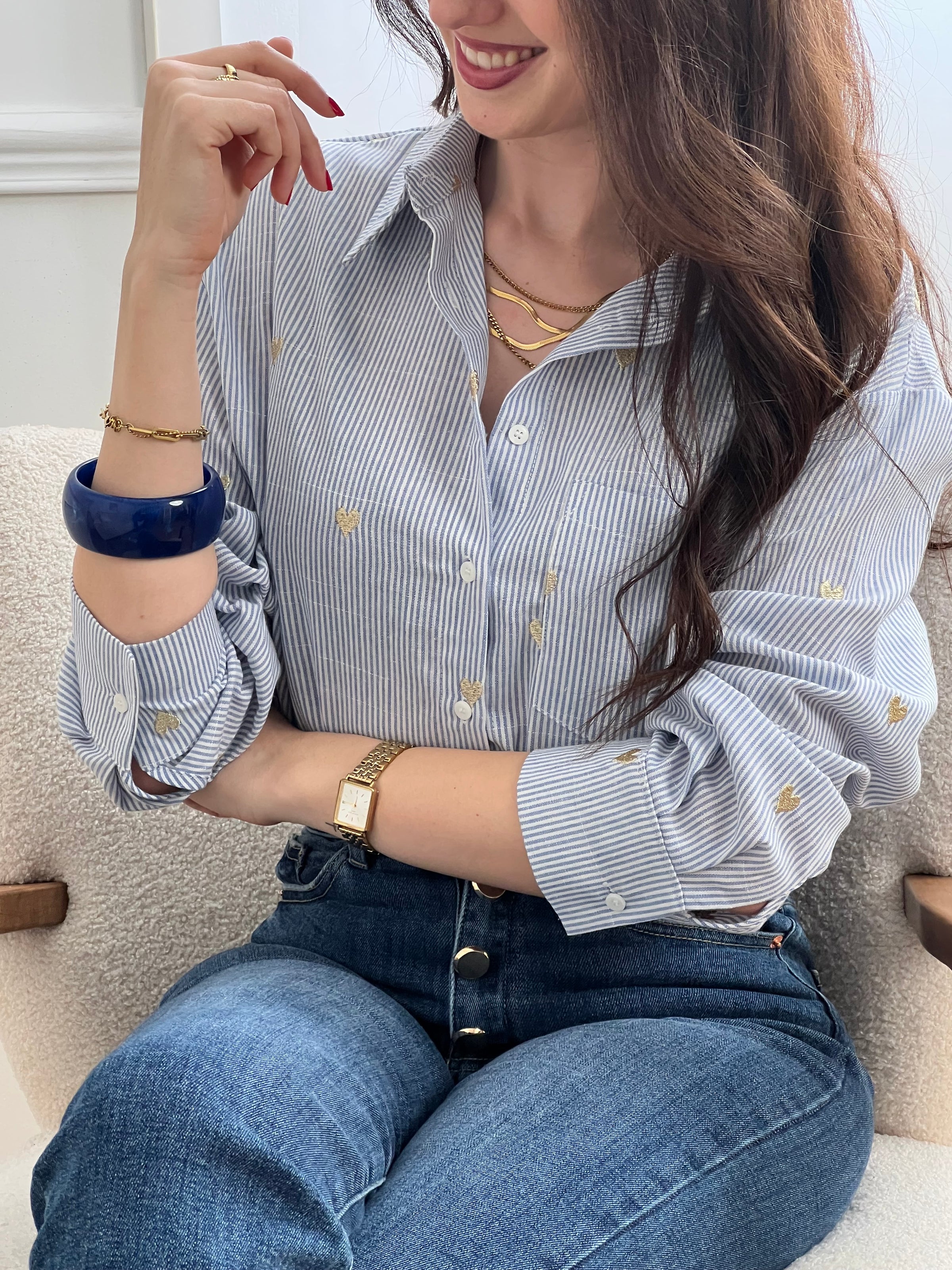 La chemise Sélène en coton combine style intemporel et détails délicats. Rayures verticales bleu et écru, cœurs dorés brodés et col chemise structuré créent une allure sophistiquée. Manches longues avec boutons aux poignets, poche poitrine et fermeture boutonnée complète.