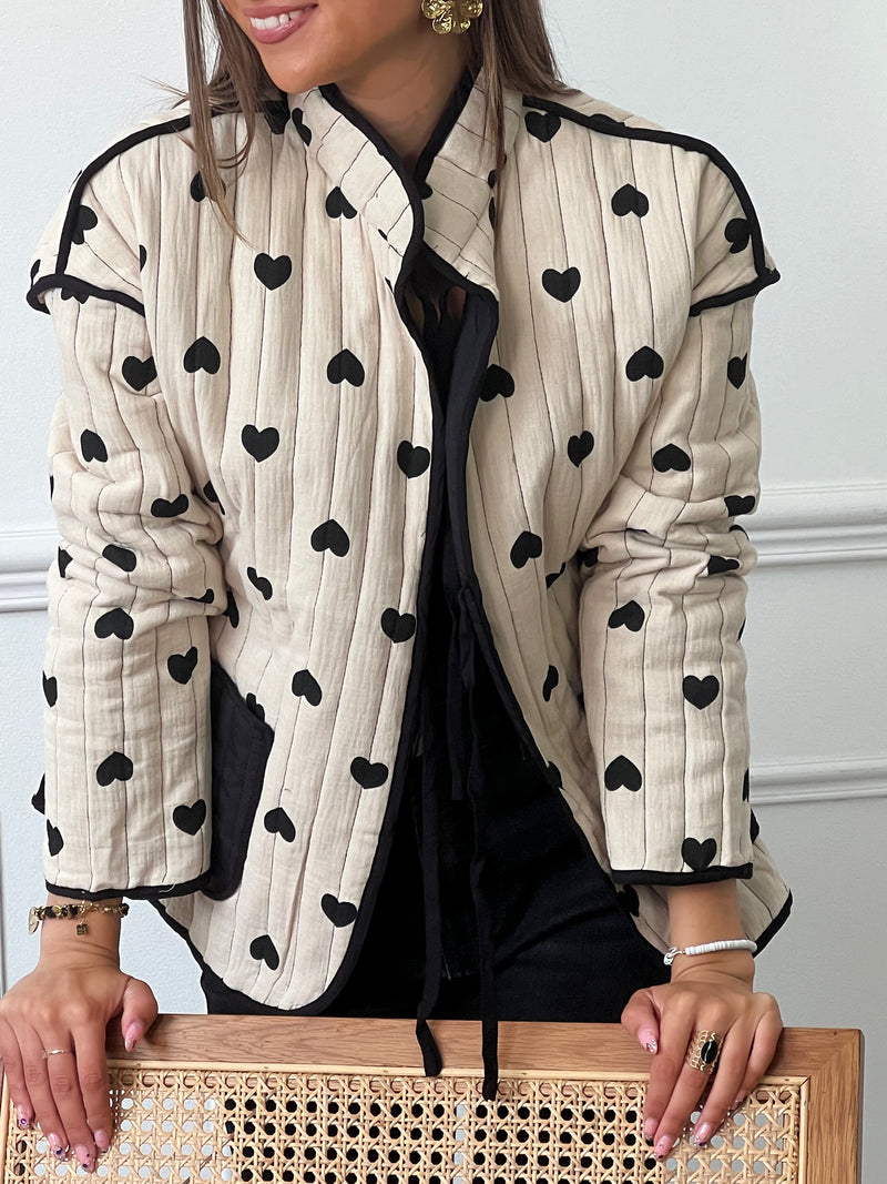 Découvrez notre veste matelassée en coton beige et noir "Joelie" avec des motifs coeurs noirs. Pratique avec ses 2 poches à l'avant, elle vous gardera au chaud avec ses manches longues et ses liens de serrage à l'avant. Ajoutez une touche de style à votre tenue grâce à ses couleurs intemporelles.