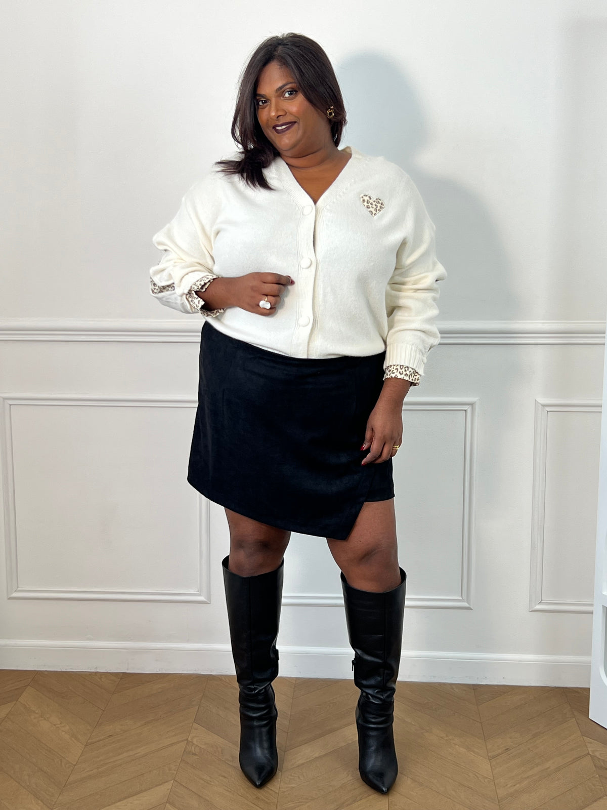Jupe-short noire Adele grande taille portée de face, avec panneau avant asymétrique, gilet écru et bottes noires.