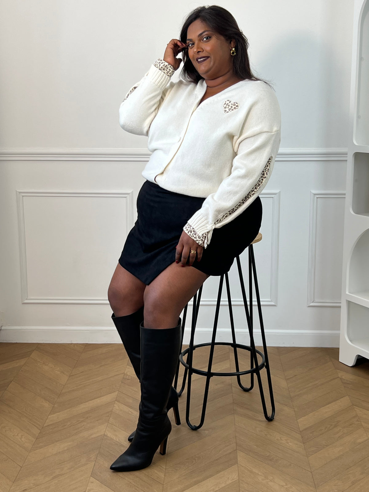 Jupe-short noire Adele portée en position assise sur un tabouret, avec un gilet écru et des bottes noires.
