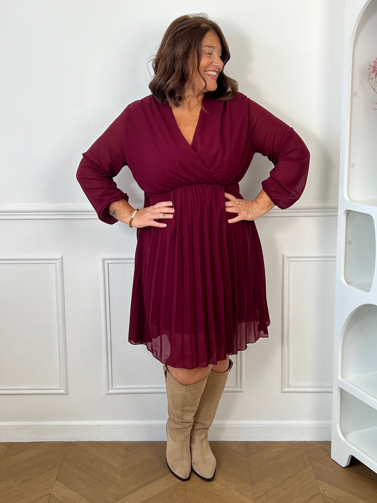 Robe courte bordeaux Ophelie grande taille portée de face, avec décolleté cache-cœur, jupe plissée et bottes beiges.