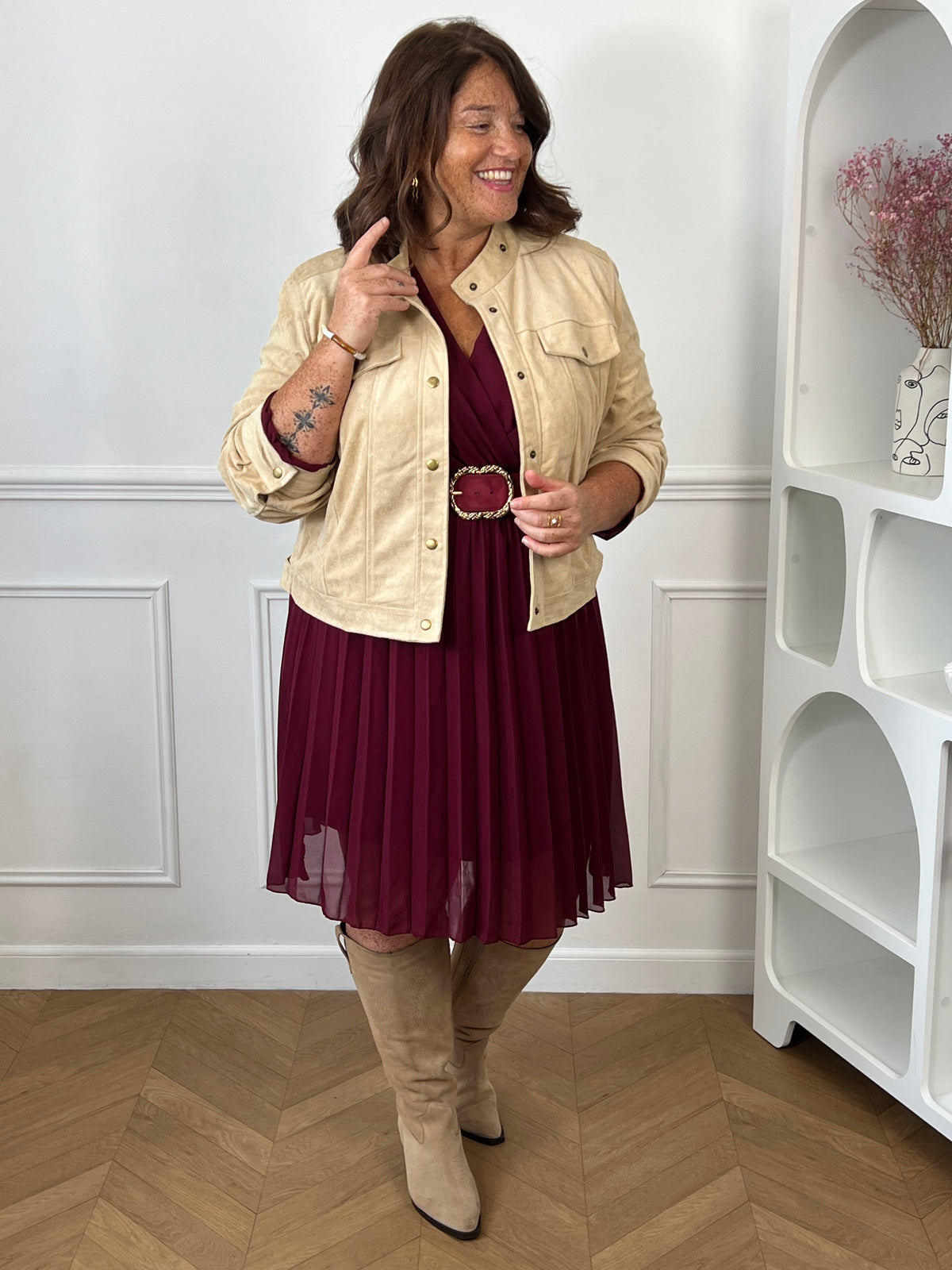 Robe bordeaux Ophelie portée avec une veste beige, une ceinture à boucle dorée et des bottes beiges.