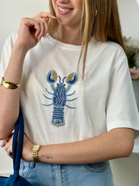 Tee-shirt écru femme en 100% coton, parfait pour un look décontracté et tendance. Avec ses manches courtes et son col rond, il offre un confort idéal au quotidien. Son motif homard en perles sur le devant apporte une touche originale et élégante. Facile à associer, ce tee-shirt aux détails écru, bleu et doré est idéal pour une tenue estivale chic et moderne.
