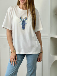 Tee-shirt écru femme en 100% coton, parfait pour un look décontracté et tendance. Avec ses manches courtes et son col rond, il offre un confort idéal au quotidien. Son motif homard en perles sur le devant apporte une touche originale et élégante. Facile à associer, ce tee-shirt aux détails écru, bleu et doré est idéal pour une tenue estivale chic et moderne.