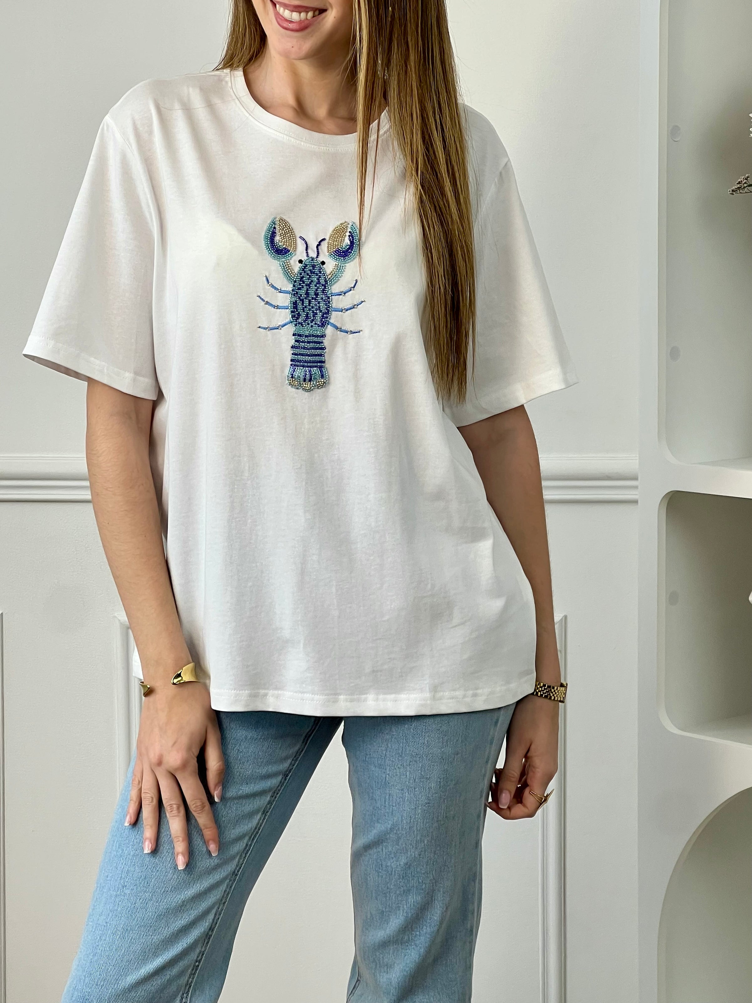 Tee-shirt écru femme en 100% coton, parfait pour un look décontracté et tendance. Avec ses manches courtes et son col rond, il offre un confort idéal au quotidien. Son motif homard en perles sur le devant apporte une touche originale et élégante. Facile à associer, ce tee-shirt aux détails écru, bleu et doré est idéal pour une tenue estivale chic et moderne.
